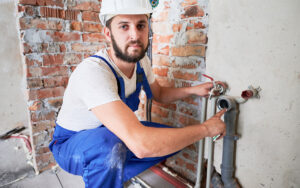 Handsome male worker in safety helmet looking at camera while applying silicone grease on pipe. Man plumber in work overalls lubricating pipe while installing water system in apartment.